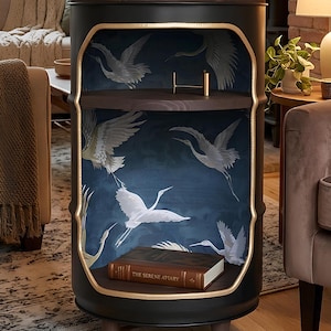 Puede incluir: Un mueble cilíndrico negro con frente abierto y borde dorado, que presenta un papel tapiz interior azul oscuro con ilustraciones de grullas blancas. La unidad tiene dos estantes de madera oscura, patas cortas y un libro titulado The Serene Apiary.
