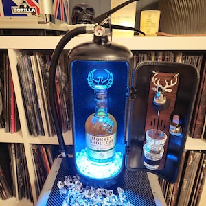 Pode incluir: Um extintor de incêndio preto reutilizado como um bar com uma prateleira de vidro, um copo de shot e uma garrafa de whisky escocês Monkey Shoulder. O bar tem uma luz azul no interior e um painel de madeira com uma silhueta de cabeça de veado.