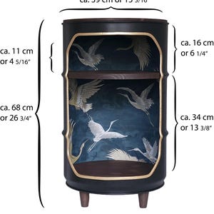 May include: A black cylindrical side table with two dark wood shelves and gold-trimmed openings. The interior features a dark blue background with white crane illustrations. Dimensions are 39 cm wide and 68 cm high, with specific shelf heights of 16 cm and 34 cm.