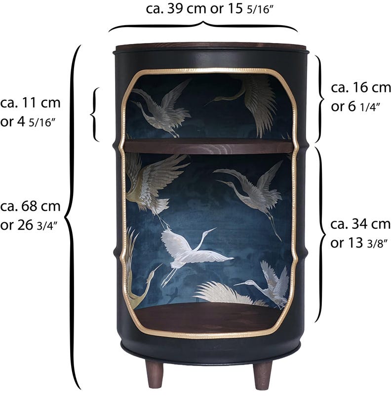 May include: A black cylindrical side table with two dark wood shelves and gold-trimmed openings. The interior features a dark blue background with white crane illustrations. Dimensions are 39 cm wide and 68 cm high, with specific shelf heights of 16 cm and 34 cm.