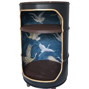 May include: A cylindrical black metal cabinet with a cutout front, featuring a dark blue interior wallpaper with white crane bird patterns. It includes two circular dark wood shelves and stands on three small, dark brown wooden legs.
