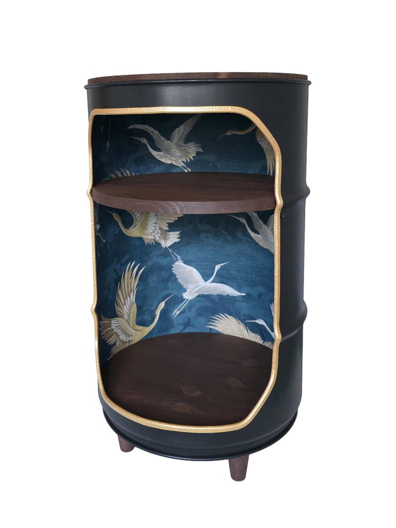 May include: A cylindrical black metal cabinet with a cutout front, featuring a dark blue interior wallpaper with white crane bird patterns. It includes two circular dark wood shelves and stands on three small, dark brown wooden legs.