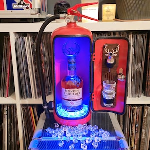 May include: A red fire extinguisher converted into a mini bar with a bottle of Monkey Shoulder blended malt scotch whisky, a shot glass, and ice cubes. The bar has a wooden door with a stag head design.