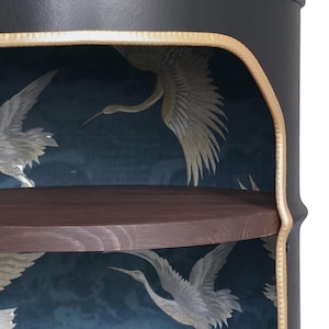May include: A cylindrical black metal cabinet with a dark wood top shelf. The interior features a deep blue wallpaper backing with a repeating pattern of white cranes in flight. The front opening is trimmed with a textured gold-coloured metal edge.