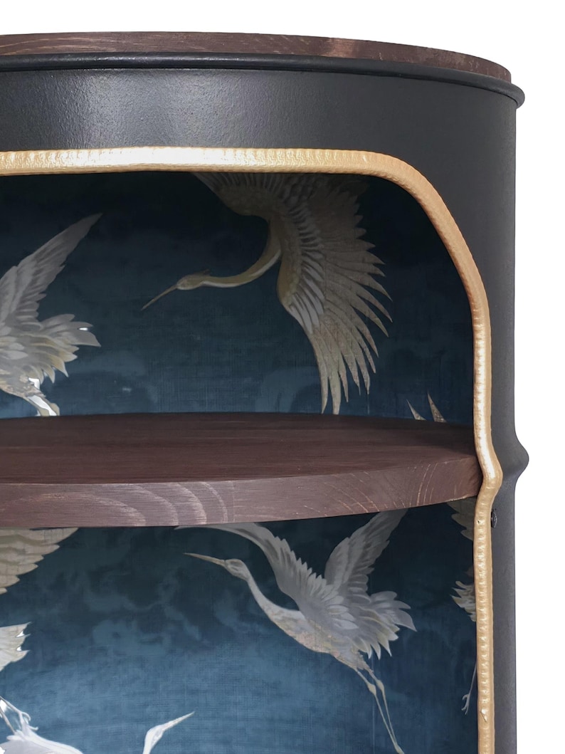 May include: A cylindrical black metal cabinet with a dark wood top shelf. The interior features a deep blue wallpaper backing with a repeating pattern of white cranes in flight. The front opening is trimmed with a textured gold-coloured metal edge.