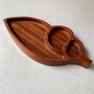 Puede incluir: Una bandeja de madera para servir con forma de hoja. La bandeja de madera de color marrón rojizo tiene tres secciones: una zona larga y curvada y dos compartimentos circulares más pequeños. Ideal para servir aperitivos.