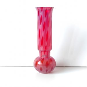 May include: A tall glass vase featuring a bulbous base and a cylindrical neck with a flared rim. The glass is translucent with a repeating pattern of vibrant red oval spots set against a soft, milky white background throughout the entire piece.