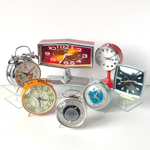 May include: A collection of vintage alarm clocks in various shapes, sizes, and colors. The clocks include a silver bell-top clock, an orange clock with a green face, and a red and silver clock. The clocks are displayed on glass shelves.