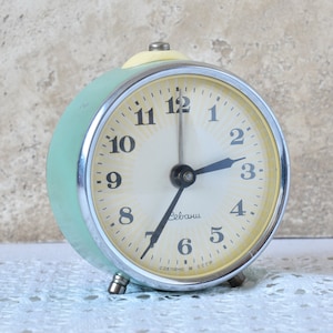 Può includere: Una sveglia vintage verde con un quadrante bianco e lancette nere. La sveglia ha una cassa cromata e una manopola gialla in cima. Il quadrante dell'orologio recita "Сделано в СССР", che si traduce in "Made in URSS".