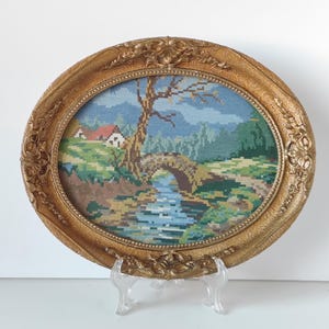 Puede incluir: Arte de bordado enmarcado ovalado que representa un paisaje con un puente, un río y una casa. El marco dorado tiene detalles ornamentados. La obra de arte presenta una paleta de colores azules, verdes, marrones y beige.