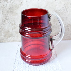 May include: A red glass beer stein with a clear glass handle. The stein has a textured, ribbed design.