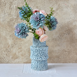 Pode incluir: Um vaso de cerâmica azul e branco com uma superfície texturizada, contendo um buquê de flores rosa e azuis. O vaso tem um design único e em camadas.