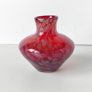Può includere: Un vaso di vetro rosso vibrante con un corpo bulboso e un collo svasato. Il vaso presenta un motivo maculato in tonalità di rosso e viola, creando un design unico e artistico. Questo vaso decorativo è adatto per esporre fiori.