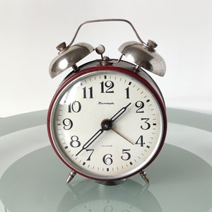 Può includere: Un orologio da sveglia vintage rosso con campane argentate e un quadrante bianco. Il quadrante dell'orologio ha indici orari in numeri romani neri e lancette nere. Il quadrante dell'orologio ha anche la scritta "Полет".