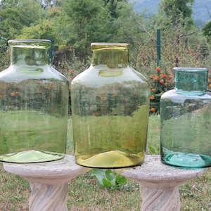 Op de afbeelding: Drie glazen potten in verschillende maten en kleuren. De grootste pot is groen, de middelste pot is geel en de kleinste pot is blauw. De potten staan op een wit voetstuk.