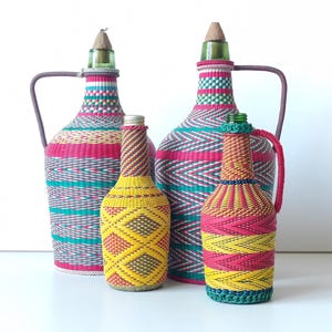 Puede incluir: Colección de botellas decorativas con exteriores tejidos en colores vibrantes. Las botellas varían en tamaño y presentan asas, tapones y patrones intrincados en rosa, turquesa, amarillo y blanco. Las botellas más grandes miden unos 30 cm de alto.