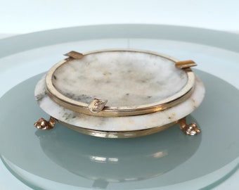 May include: A white marble ashtray with a gold rim and three gold feet. The ashtray is round and has a slight indentation in the center.