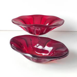 Può includere: Due ciotole di vetro rosso rubino con un bordo ondulato a forma di petalo. Le ciotole sono impilate, una leggermente sopra l'altra, e riflettono la luce, mostrando la loro finitura lucida. Le ciotole sono un oggetto decorativo.