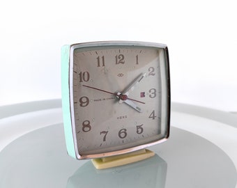 May include: A vintage, square-shaped, mint green alarm clock with a white face and black numbers. The clock has a silver rim and a white base. The clock face reads "MADE IN CHINA" and "中国製".