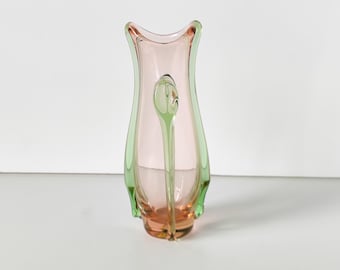 May include: A decorative glass vase with a unique design. The vase has a soft pink interior and is outlined with a light green border. A green, elongated glass element is inside the vase. The vase is a decorative home accent.
