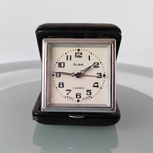 Könnte beinhalten: Ein quadratischer Vintage-Reisewecker mit cremefarbenem Zifferblatt, schwarzen Ziffern und dem Markennamen "SLAVA". Die Uhr ist in einem schwarzen Kunstleder-Etui untergebracht. Die Worte "11 JEWELS" und "MADE IN USSR" sind auf dem Zifferblatt aufgedruckt.