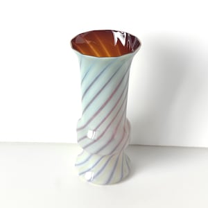 May include: A decorative glass vase with a twisted design. The vase features a light blue and white spiral pattern with red and purple stripes. The interior of the vase is a warm orange color.