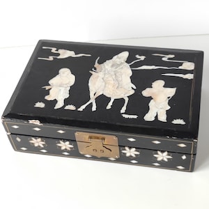 Peut inclure: Boîte en bois noir avec un couvercle à charnières décoré d'incrustations de nacre blanche représentant des personnages et un cheval. La boîte est munie d'un loquet en laiton.