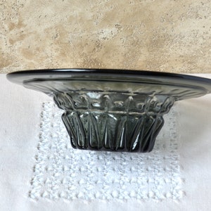 May include: A dark gray glass bowl with a wide, flat base and a fluted, cylindrical center. The bowl is sitting on a white lace doily.