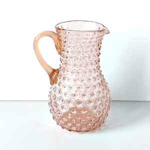 May include: A translucent light pink glass pitcher featuring a textured hobnail pattern across its rounded body. The vessel includes a curved handle on the side and a small pouring spout at the top rim, showcasing a consistent pale rose hue.