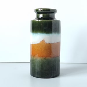 Op de afbeelding: Een cilindervormige keramische vaas met een glanzende afwerking. De vaas heeft horizontale kleurstroken: donkergroen aan de boven- en onderkant, met een witte en oranje band in het midden. De vaas heeft een smalle hals en een licht uitlopende rand.