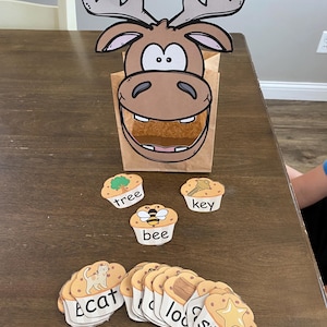 May include: A brown paper bag decorated with a cartoon moose head, with a cutout mouth. The bag is filled with a brown, textured material. Several word cards and cupcake-shaped cards with words and images, including "cat", "bee", and "star", are scattered around.
