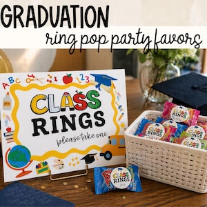 Op de afbeelding: Een display met een diploma-uitreikingsthema met een bord met de tekst "CLASS RINGS, please take one." Een witte mand bevat ring pop snoepjes. Het bord bevat schoolgerelateerde illustraties en de tekst "GRADUATION ring pop party favors."