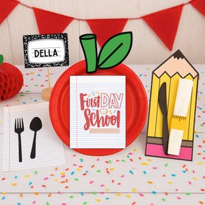 May include: A festive back-to-school themed table setting. A red plate is topped with a notebook-shaped card that reads "First Day of School." A pencil-shaped holder contains a knife and two white erasers. A notebook place card reads "DELLA."