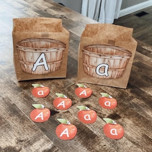May include: Two brown paper bags with a basket illustration and the letter "A". Scattered around are red apple-shaped cutouts, each with the letter "a" or "A" and a green leaf. The items are on a wooden surface.