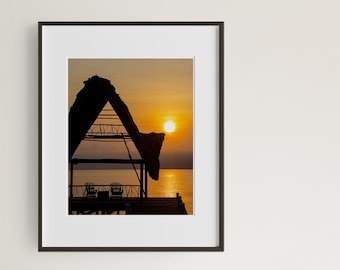 May include: Silhouette of a thatched roof hut on a wooden pier overlooking a body of water at sunset. The sun is setting behind the hut, casting a warm glow on the water.