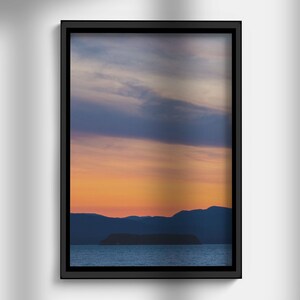 May include: A framed photograph depicting a sunset over a body of water, with a small island in the foreground. The sky is a blend of orange, pink, and purple shades, with clouds casting long shadows on the water.