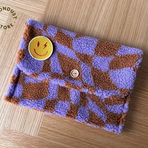 Op de afbeelding: Een pluche clutch met een geruit patroon in paars en bruin, met een gele smiley-patch en een bruine knoop.