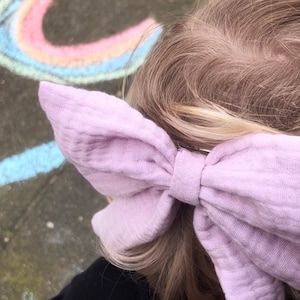 Op de afbeelding: Een grote, lichtroze strik van stof, gebonden in het haar van een persoon met blond haar. De strik is gemaakt van een zachte, textuurrijke stof.