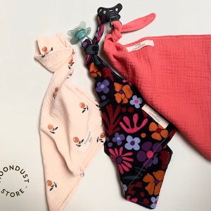 Op de afbeelding: Drie baby-slabbetjes met verschillende patronen. Eén is roze met een klein oranje printje, één is koraal en één is zwart met een kleurrijke bloemenprint. Elk slabbetje heeft een wit label met de tekst "MOONDUST STORE".