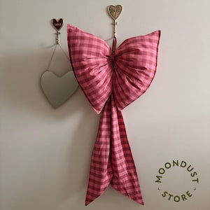 Op de afbeelding: Een grote, roze ruitjesstrik hangt aan een witte muur. De strik is gemaakt van een zachte stof en heeft lange, vloeiende uiteinden. Twee hartvormige haken en een hartvormige spiegel zijn ook zichtbaar. De tekst "MOONDUST STORE" staat in een cirkel.