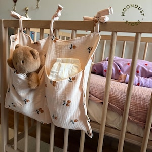 Op de afbeelding: Een perzikroze bedorganizer met een kersenpatroon, met een teddybeer en luiers. De organizer is bevestigd aan een houten ledikant. De afbeelding toont ook een slapende baby in een roze en paarse outfit. Het Moondust Store-logo is zichtbaar.