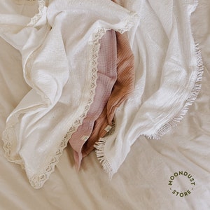 May include: Two white muslin swaddles with lace trim and fringe. One swaddle is folded and the other is draped over it. The swaddles are on a light brown surface.