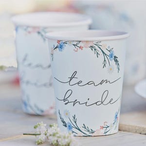 Könnte beinhalten: Zwei weiße Pappbecher mit floralen Designs und dem Schriftzug "team bride" in Schreibschrift. Die Becher haben einen hellblauen Hintergrund mit einem Kranz aus rosa, blauen und weißen Blumen. Ideal für eine Brautparty.