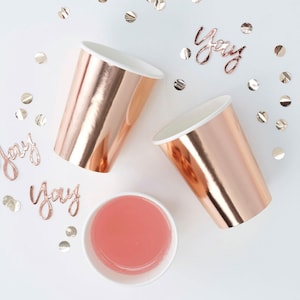 Könnte beinhalten: Zwei roségoldene Pappbecher mit weißen Rändern und ein Becher mit rosa Flüssigkeit auf einer weißen Oberfläche, übersät mit silbernen Konfetti. Das Wort "Yay" ist aus roségoldenem Draht geschrieben. Partyzubehör.