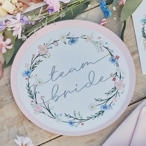 Könnte beinhalten: Ein runder Pappteller mit rosa Rand und floralem Kranzdesign. Der Kranz zeigt rosa, blaue und weiße Blumen mit grünen Blättern. Die Worte "team bride" sind in Schreibschrift in der Mitte des Tellers geschrieben.