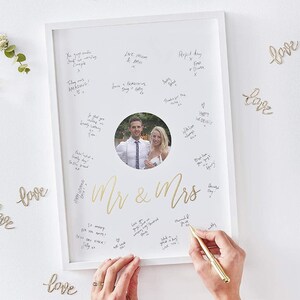 Könnte beinhalten: Ein weiß gerahmtes Hochzeitsgästebuch mit einem Foto eines Paares in der Mitte. Die Worte "Mr & Mrs" stehen in goldener Schrift unter dem Foto. Gäste haben Nachrichten um das Foto herum mit einem goldenen Stift geschrieben.