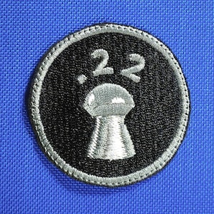 May include: Embroidered patch with a black circular design and a silver border. The patch features the number ".22" above a silver keyhole. The background is a vibrant blue.