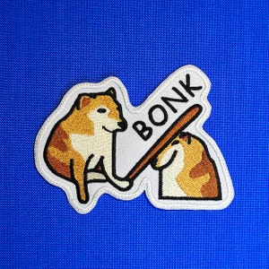 Può includere: Toppa ricamata con due cani dei cartoni animati, uno con una mazza da baseball e l'altro che viene colpito. La parola "BONK" è scritta in maiuscolo nero su uno striscione bianco. La toppa ha un bordo bianco su sfondo blu.