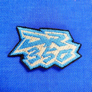 May include: Embroidered patch with the text "ZR 350" in light blue and beige, outlined in black, on a blue fabric background. The patch has a unique, angular shape.