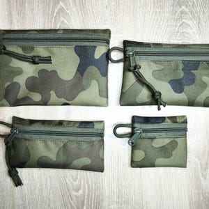 Peut inclure: Quatre pochettes zippées de camouflage vert olive de différentes tailles. Chaque pochette a une fermeture éclair assortie et une petite boucle avec un cordon noué. Les pochettes sont disposées sur une surface claire imitation bois.